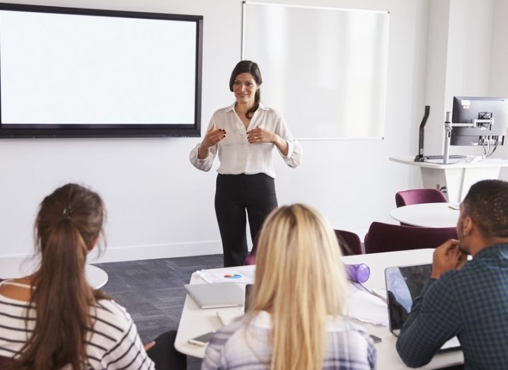 A woman giving a lecture to students in a university classroom, with a whiteboard on the wall.