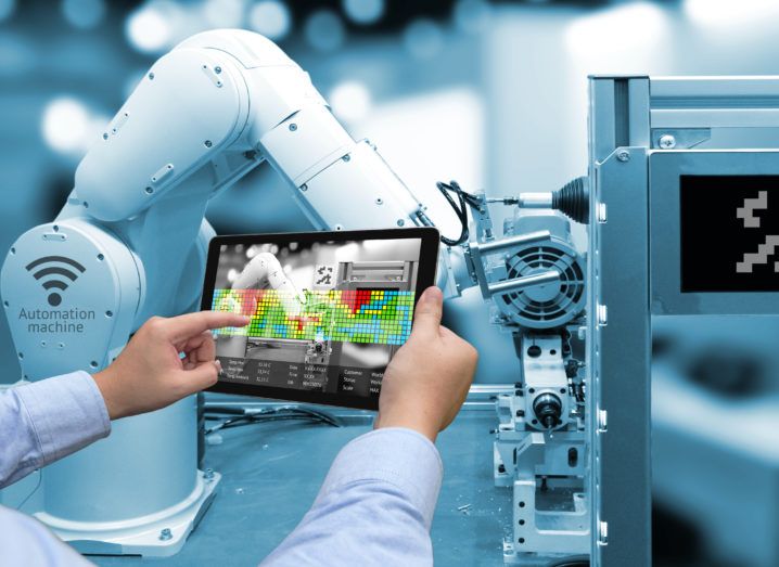 Factory worker holds up a computer tablet beside a robotic arm.