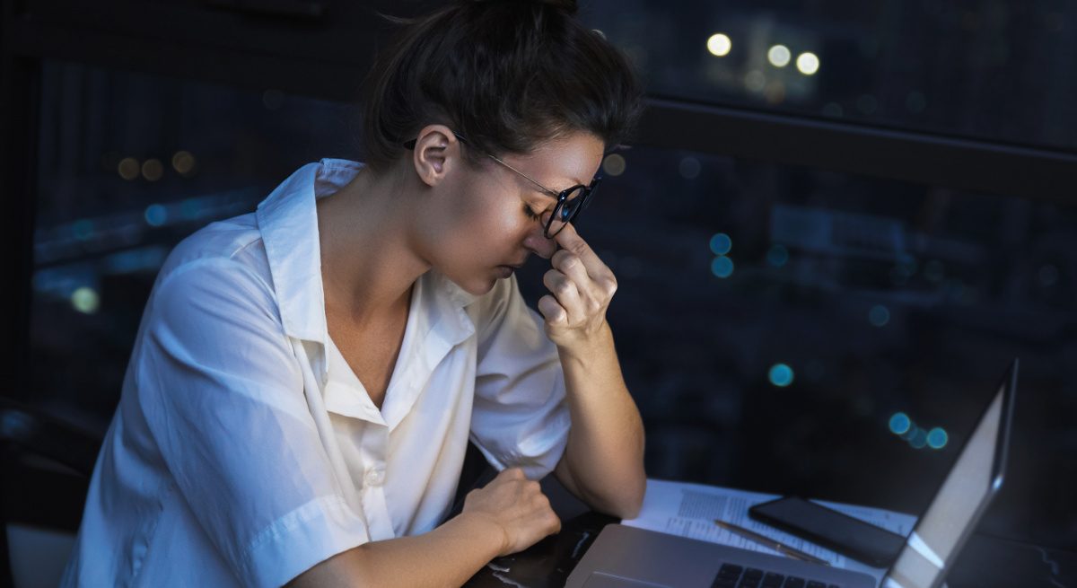 A female employee looks exhausted as she pinches her nose due to work-related stress in a dark room in front of a laptop.