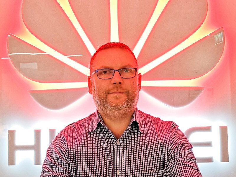 Man in check shirt and glasses sitting in front of a Huawei logo in lights.