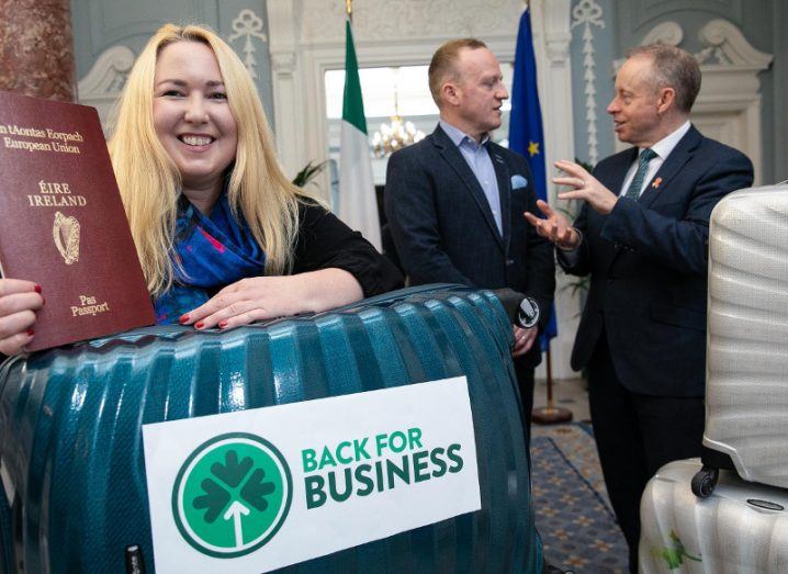 Woman with blonde hair holds an Irish passport and green suitcase while two men in suits chat in background.