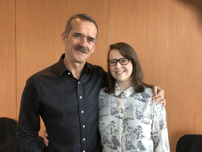middle aged man with grey moustache and dark shirt with his arm around young woman with brown hair, glasses and a white shirt with black floral print.