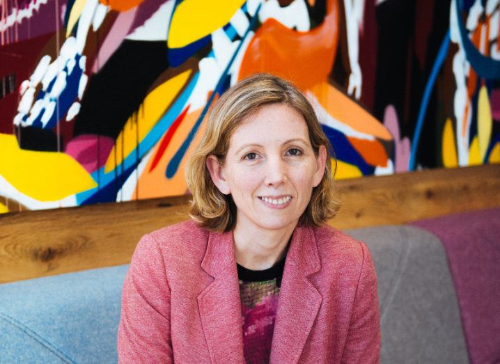 woman with short blonde curly hair wearing pink blazer and sitting on couch in front of multicoloured mural.