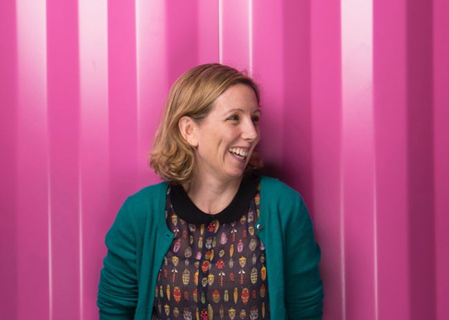 woman with short blonde curly hair wearing patterned dress and green cardigan, laughing and standing in front of bright pink wall.