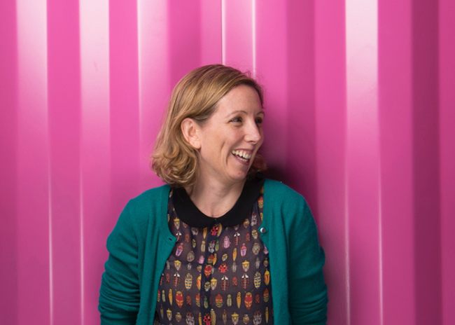 Medb Corcoran looks to the left and smiles, standing against a bright pink background.