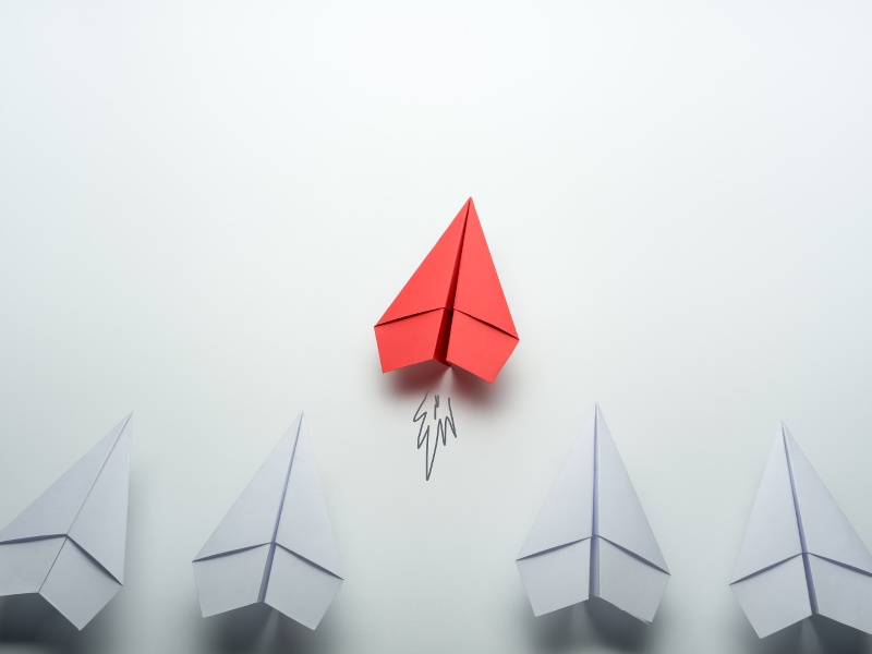 A row of white paper aeroplanes with one red one taking off.