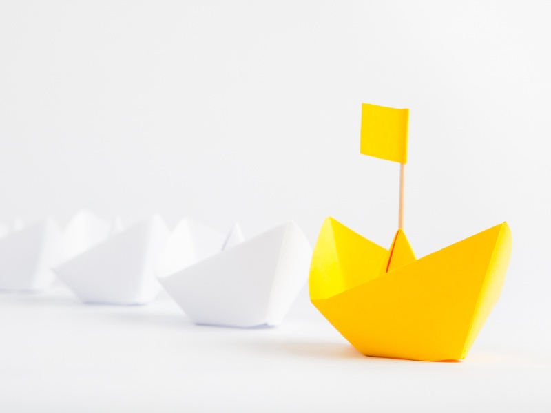 A row of white paper origami boats being led in front by a yellow boat with a yellow flag.