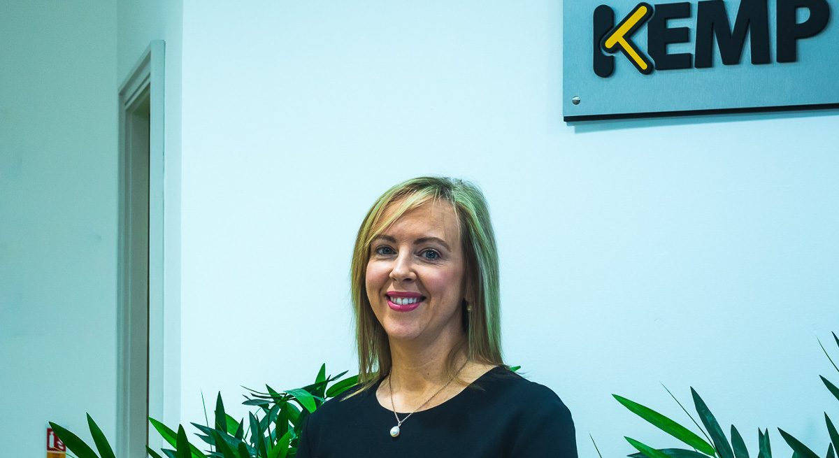 A blonde woman smiling in an office with the Kemp Technologies logo in the background on the wall.