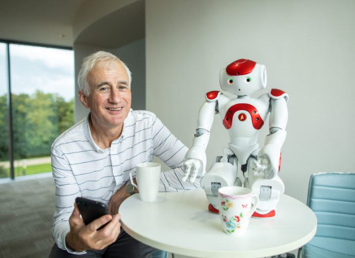 Man in stripy shirt holding smartphone beside a robot.