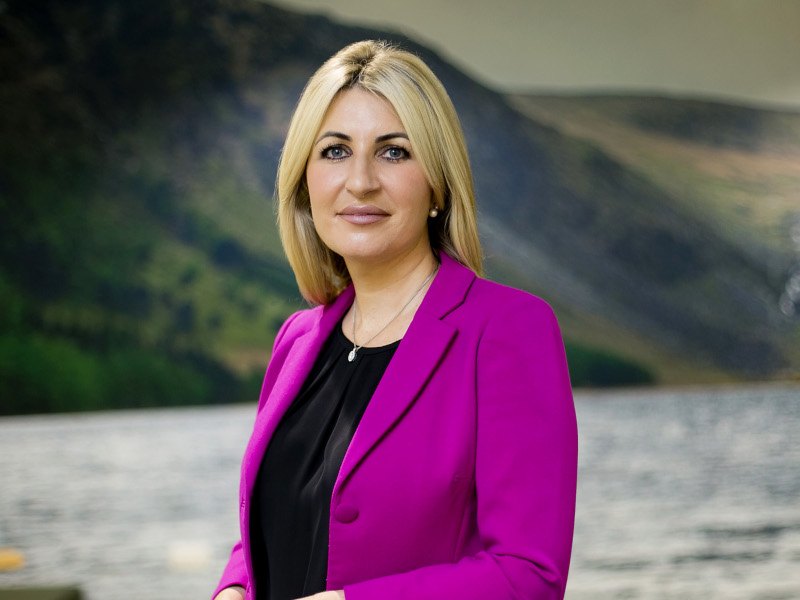 Woman with blonde hair and in pink jacket stands before a lake.