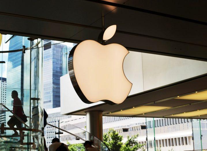 A white Apple logo suspended in the air against with a glass staircase and window in the background.