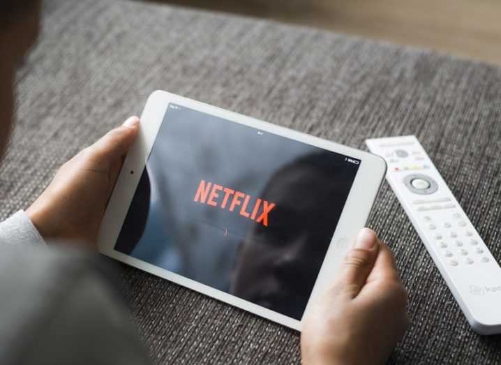 Hands holding a tablet displaying the Netflix logo , resting on a grey couch.