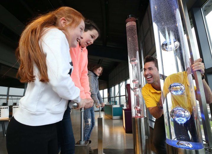 Two happy girls pressing down on a plunger forcing air bubbles into water in a tube. A man in a yellow tshirt is guiding them.