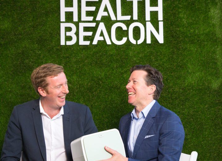 Two men in dark jackets sit before a green wall holding a white medical device in front of a green wall with HealthBeacon branding in white.