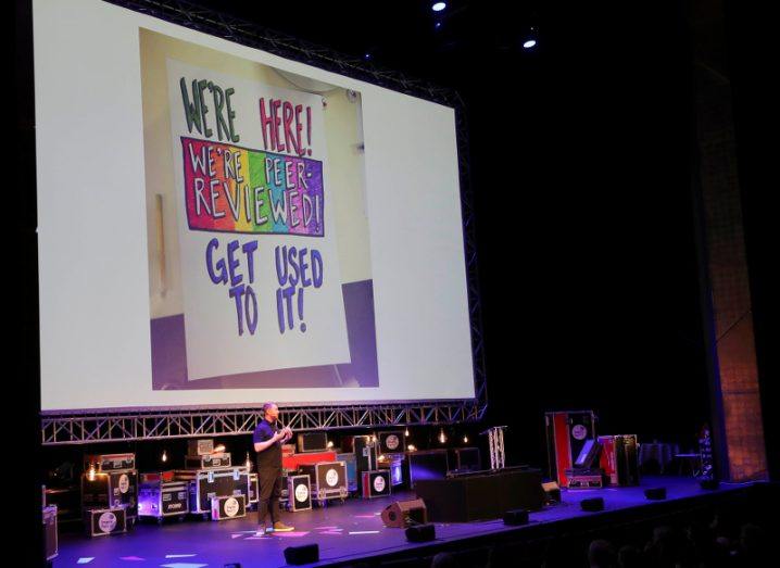 Wide view of a stage and speaker with a large background screen displaying ‘We’re here, we’re peer-reviewed, get used to it!’