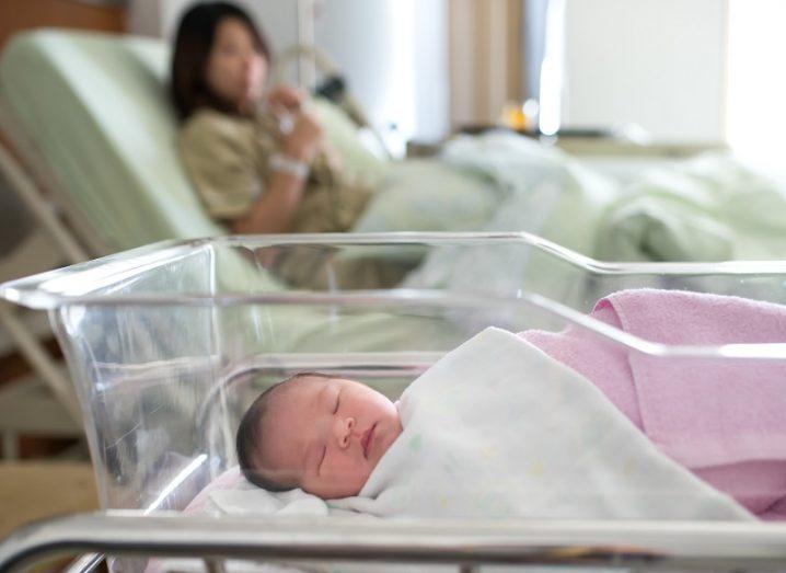 Newborn baby in a cot while their mother in hospital bed looks on in the background.