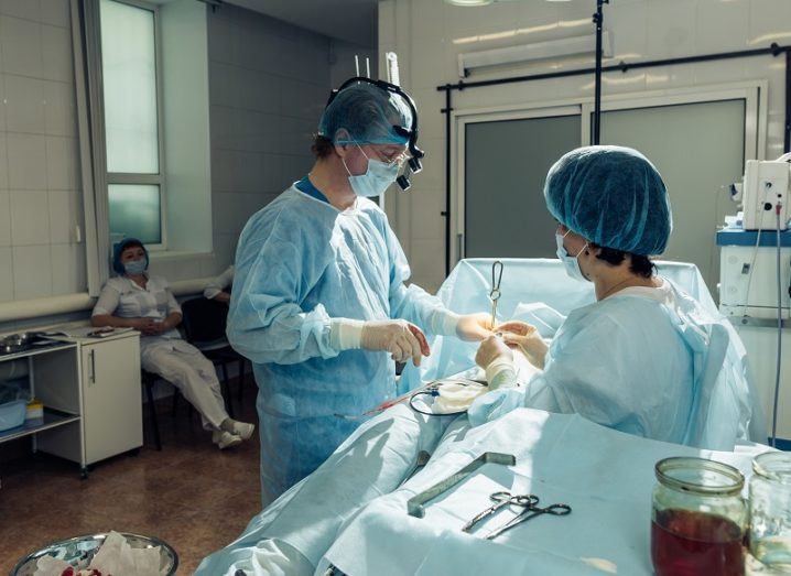 Surgeon and nurse in blue gowns operating on a patient in a dimly lit room with one spotlight.
