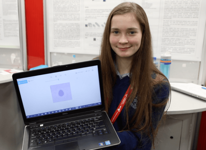 A young brown-haired girl in school uniform smiling while holding up a laptop at BT Young Scientist 2019.