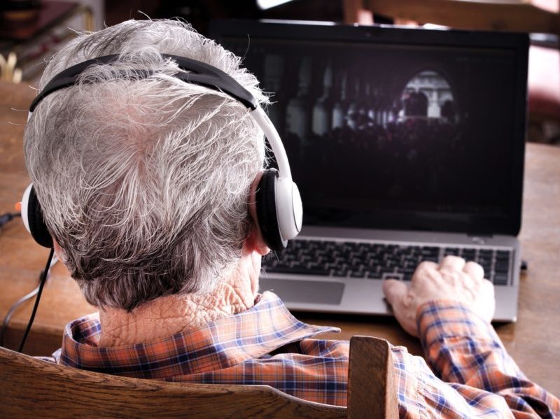 Back of an older man's head wearing headphones as he uses a laptop computer.