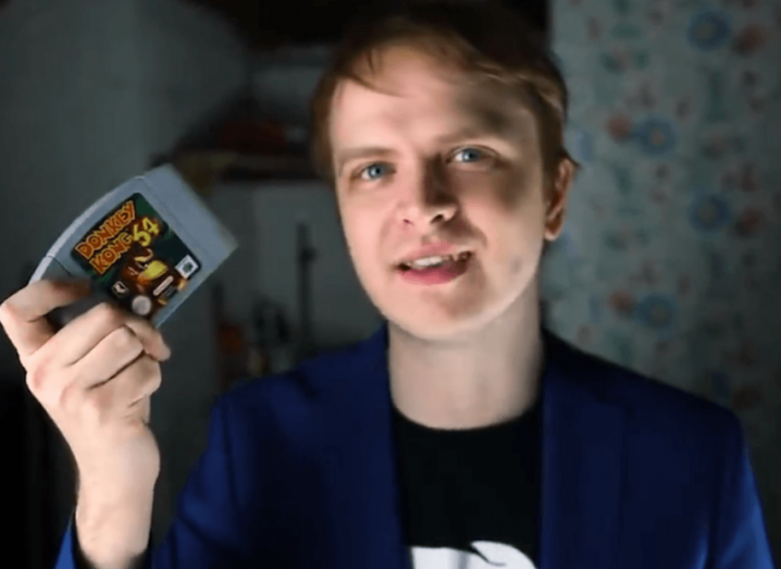 YouTuber H.Bomberguy holding a Donkey Kong 64 game cartridge and wearing a black t-shirt.