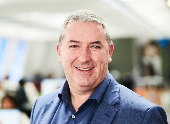 Middle-aged man with grey hair and navy suit smiling at the camera against blurred office background.