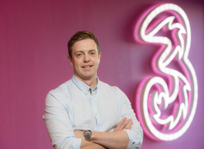 Man in white shirt stands with arms folded against a pink wall with a giant neon sign denoting Three Ireland logo.
