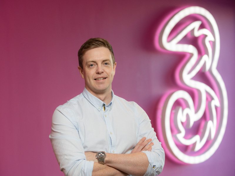 Man in white shirt stands with arms folded against a pink wall with a giant neon sign denoting Three Ireland logo.