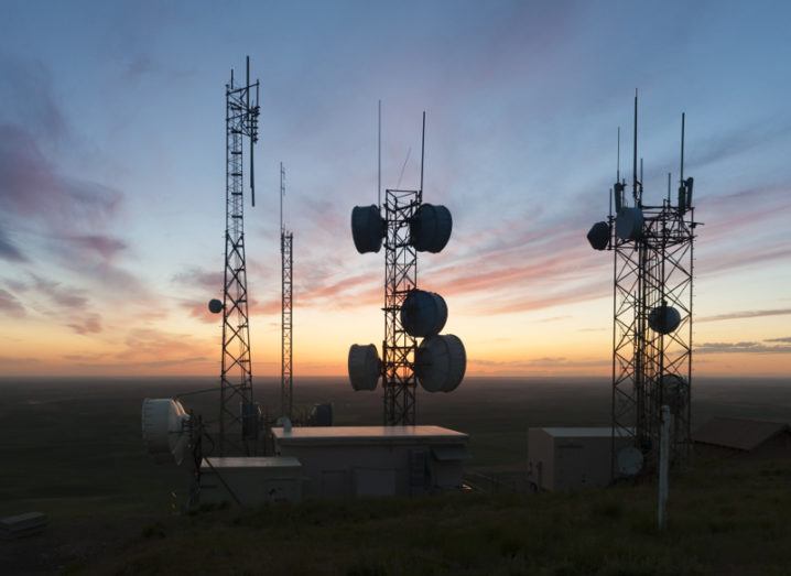 Silhouettes of radio towers on a hill against a setting sun.