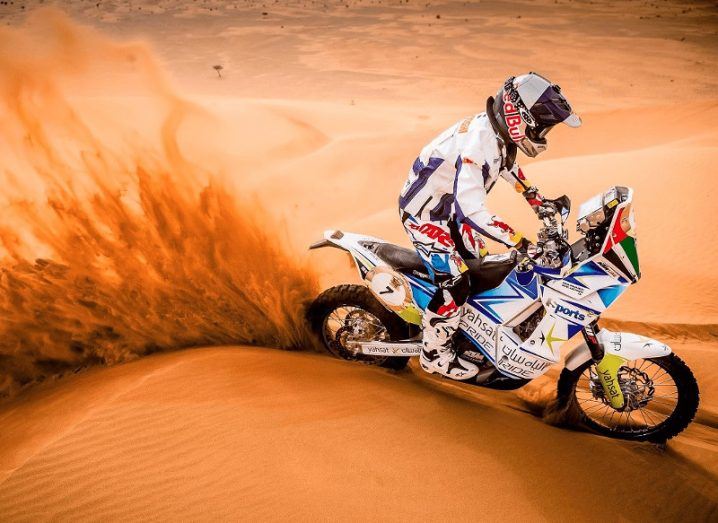 A picture of a motorcyclist driving through the desert, kicking up orange sand in his wake.