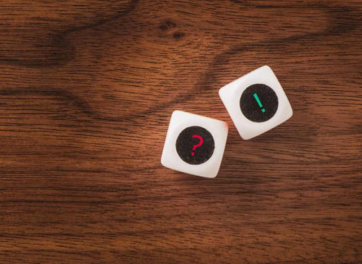 Two white dice one a wooden table, one dice showing a red question mark, the other a green exclamation mark.