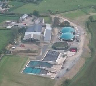 Grainy aerial image of anaerobic digestion plant, with circular blue containers visible beside a shed construction, on farm site surrounded by green fields.