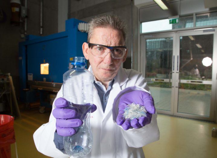 Middle-aged male researcher in white lab coat and purple gloves holds up a plastic bottle in one hand and in the other a composite raw material that can be used to make car and tractor parts.