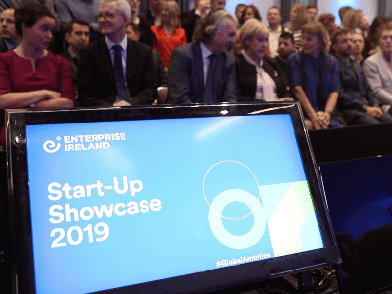 A large group of people sitting behind a blue screen that says Start-up Showcase 2019.