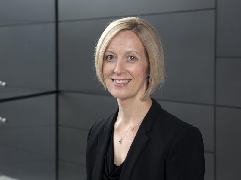 A woman with blonde hair wearing a black suit jacket standing in front of a grey wall.