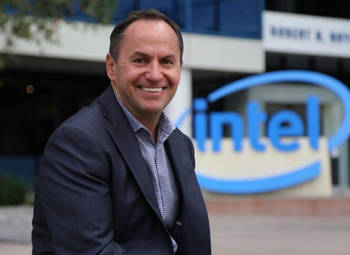 Man in dark jacket and blue shirt smiling in front of an Intel sign.