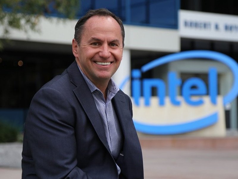 Man in dark jacket and blue shirt smiling in front of an Intel sign.