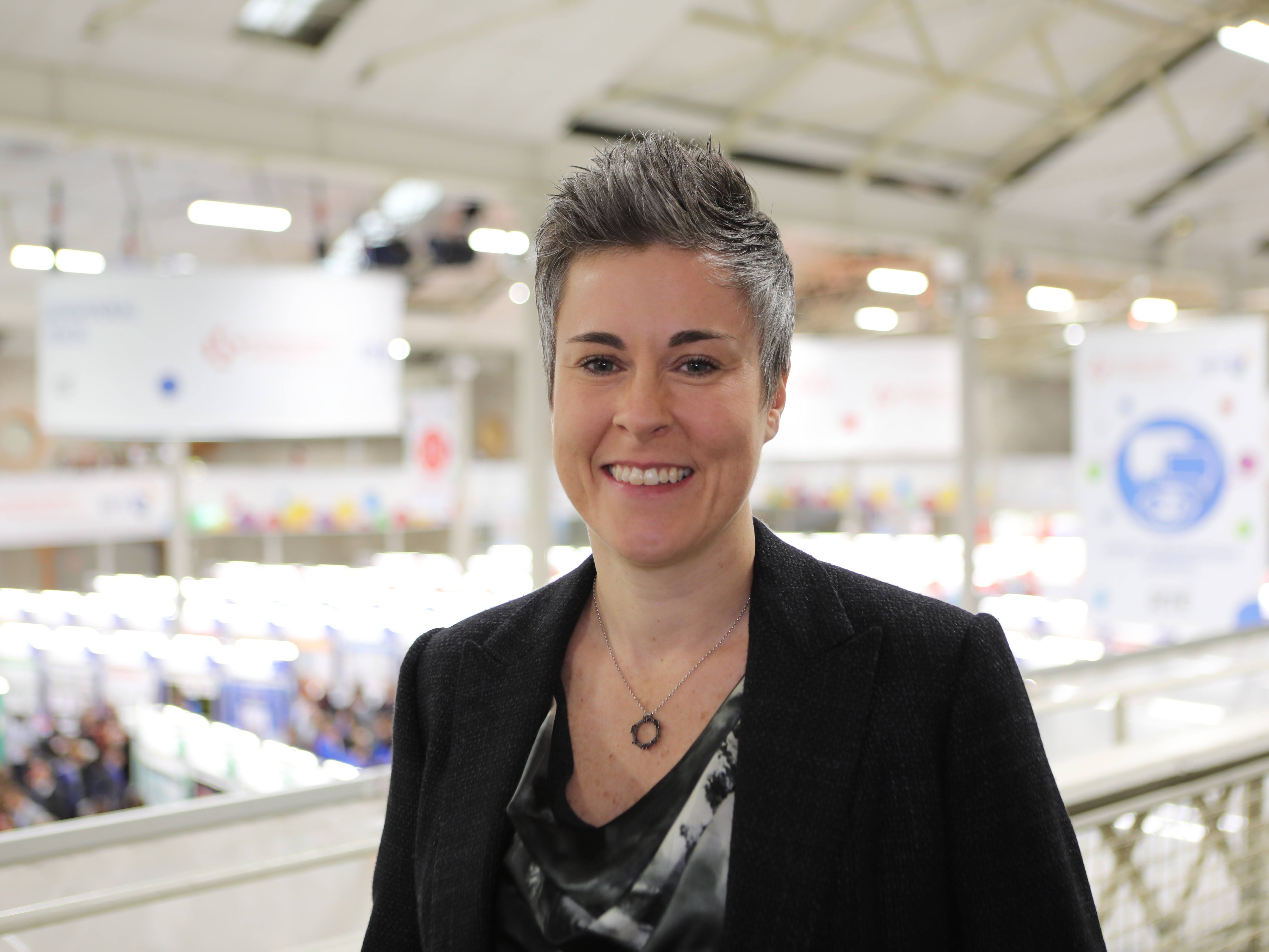 Picture of a smiling woman with short cropped hair in a black jacket at a science fair.