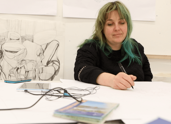A woman with dyed green hair draws something on a table next to a tablet and smartphone. Detailed line drawings hang on the wall behind her.