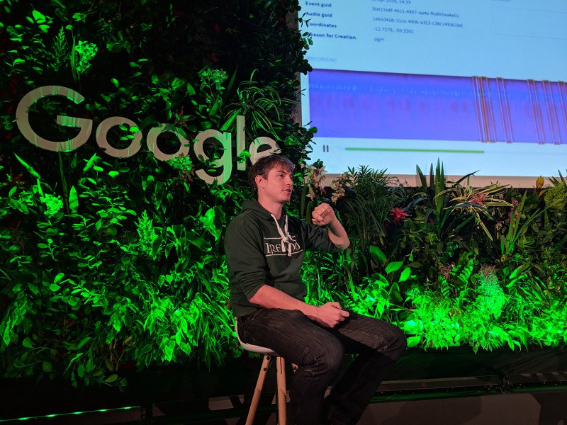 Young man in an Ireland rugby hoodie on stage at a Google event surrounded by foliage under a large screen.