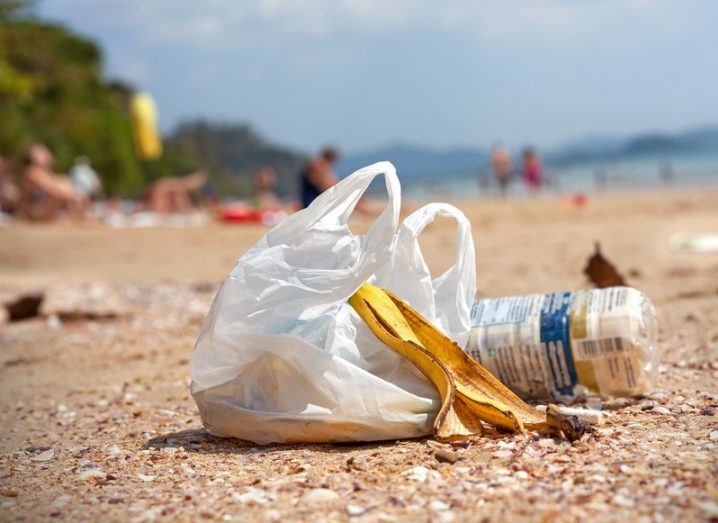 Plastic bag full of rubbish left behind on a beach, with people sitting on the sand in the background.