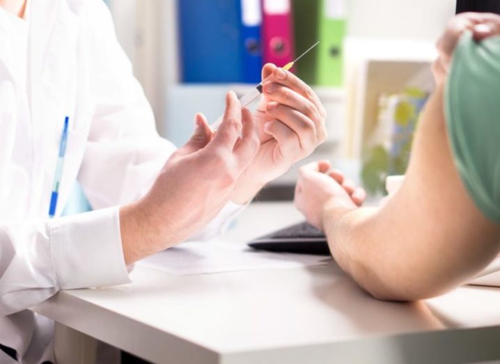 A doctor in a hospital room, wearing a white coat administering a vaccine to a patient via a needle in the upper arm.