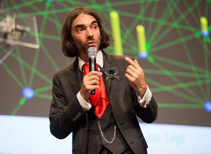 Cedric Villani giving a talk, wearing a three-piece suit, red cravat, and a brooch in the shape of a spider on the lapel.