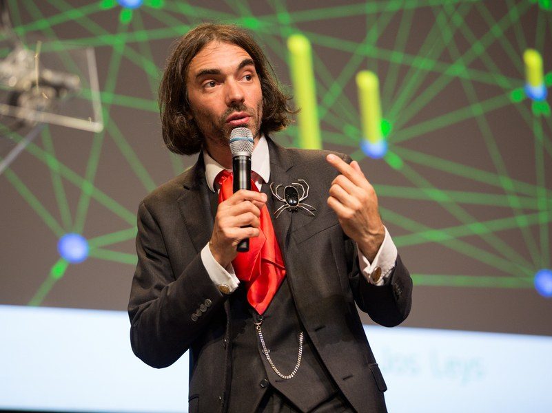 Cedric Villani giving a talk, wearing a three-piece suit, red cravat, and a brooch in the shape of a spider on the lapel.