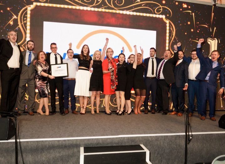 A group of diverse, formally dressed employees standing on a stage cheering at the Great Place to Work awards.