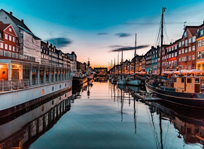 View of Nyhavn in Copenhagen at golden hour, framed by picturesque colourful houses on either side with ships and reflective water in the centre.