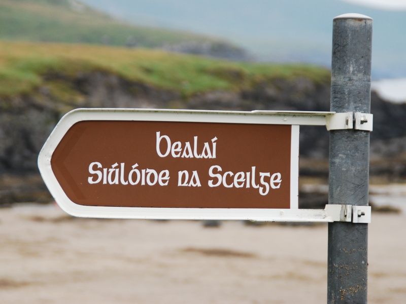 A signpost on a rocky beach reads ‘Bealaí Siúlóide na Sceilge’ in old Irish writing.