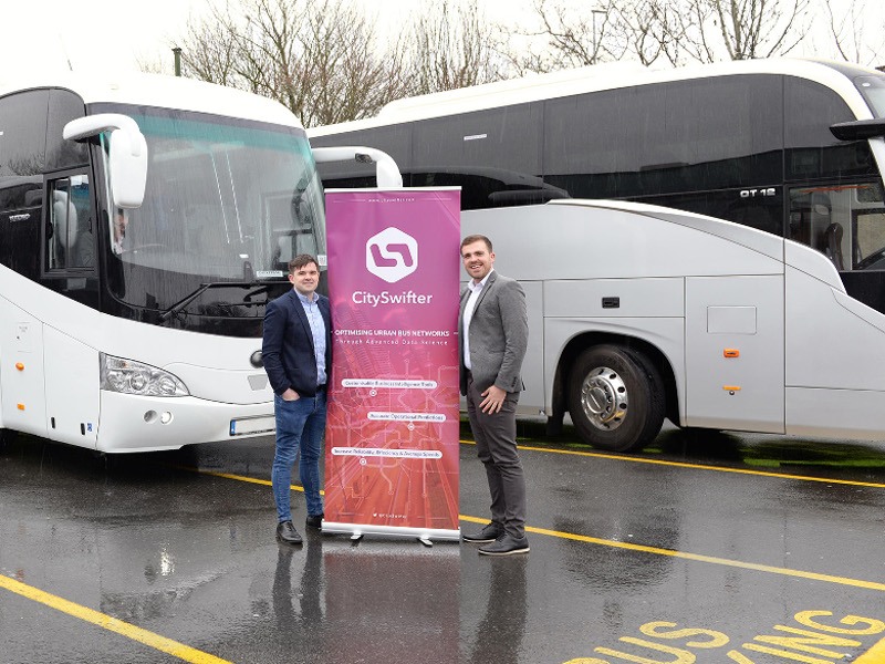 Two men hold a pink pop-up sign that reads CitySwifter in front of large buses.