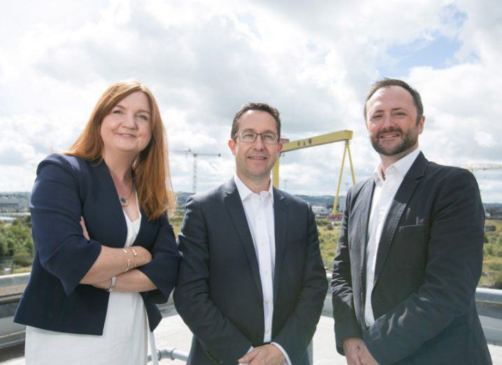 Woman with red hair and navy jacket stands beside two men in suits on a rooftop of a Belfast building with shipbuilding cranes in background.