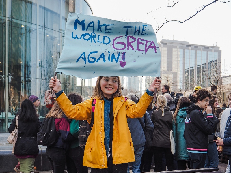 A young girl in a yellow raincoat stands on a bench amid a crowd of protesters holding up a sign that reads: ‘Make the world Greta again!’