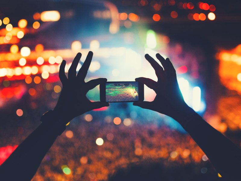 Silhouetted arms hold a smartphone up against a crowded concert lit with colourful lighting. The phone is capturing a video.